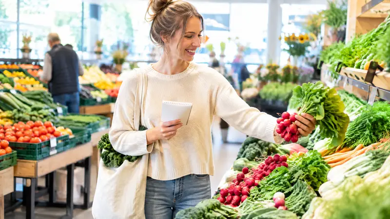 Frau mit Einkaufszettel und Stoffbeutel steht in der Abteilung für Obst und Gemüse eines Supermarkts