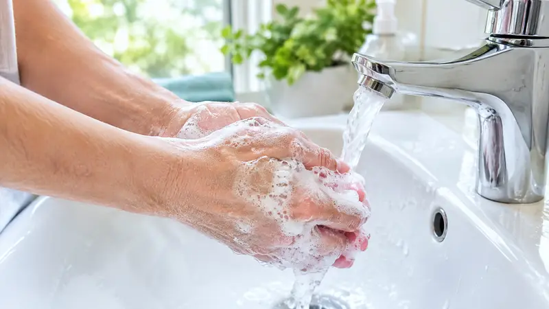 Zwei mit Seifenschaum bedeckte Hände unter einem fließenden Wasserstrahl über einem weißen Waschbecken.