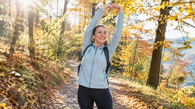 Eine Frau streckt sich gut gelaunt mit erhobenen Armen auf einem Waldweg, der mit buntem Herbstlaub bedeckt ist.