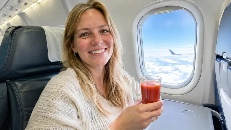 Ein Passagier sitzt am Flugzeugfenster und hält ein Glas Tomatensaft in der Hand, draußen sind Wolken zu sehen.